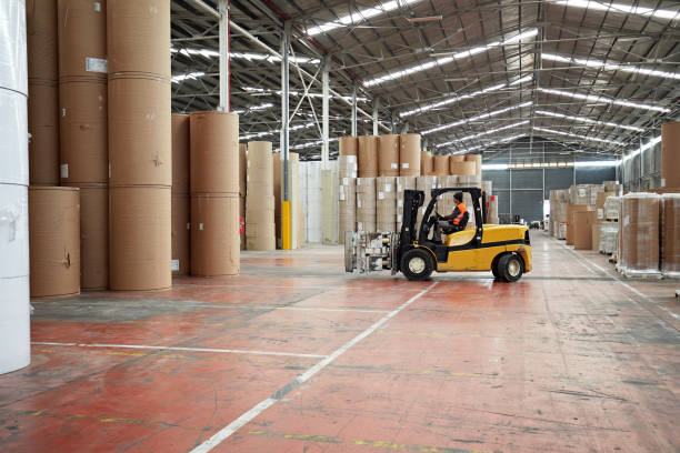Full length side view of warehouseman in early 20s wearing reflective vest over work attire and driving vehicle with paper roll clamp attachment.